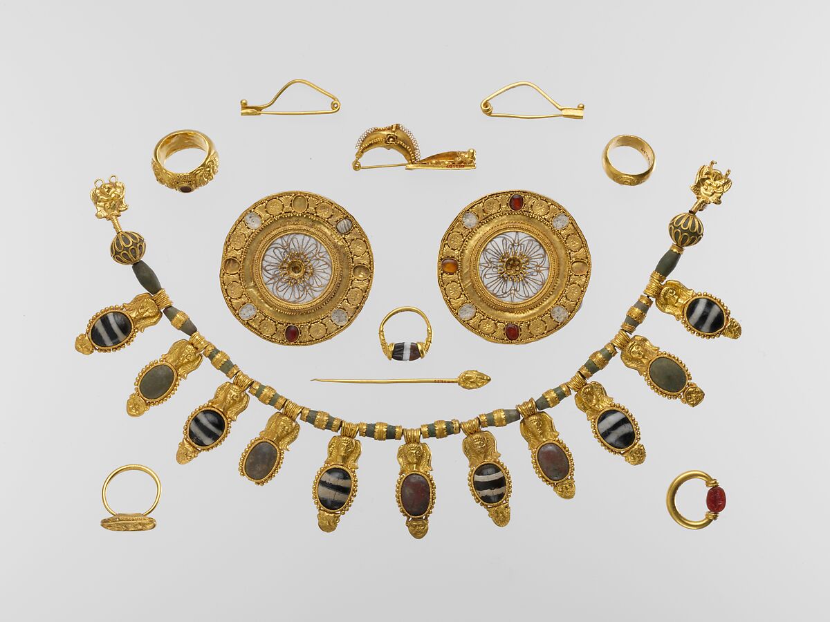 From the description of the Metropolitan Museum of Art: "The tomb group represents one of the richest and most impressive sets of Etruscan jewelry ever found. It comprises a splendid gold and glass pendant necklace, a pair of gold and rock-crystal disk earrings, a gold dress fastener (fibula) decorated with a sphinx, a pair of plain gold fibulae, a gold dress pin, and five finger rings. Two of the rings have engraved scarabs that revolve on a swivel bezel; one is decorated with embossed satyr heads, and the other two have decorated gold bezels" (MET, 2021).