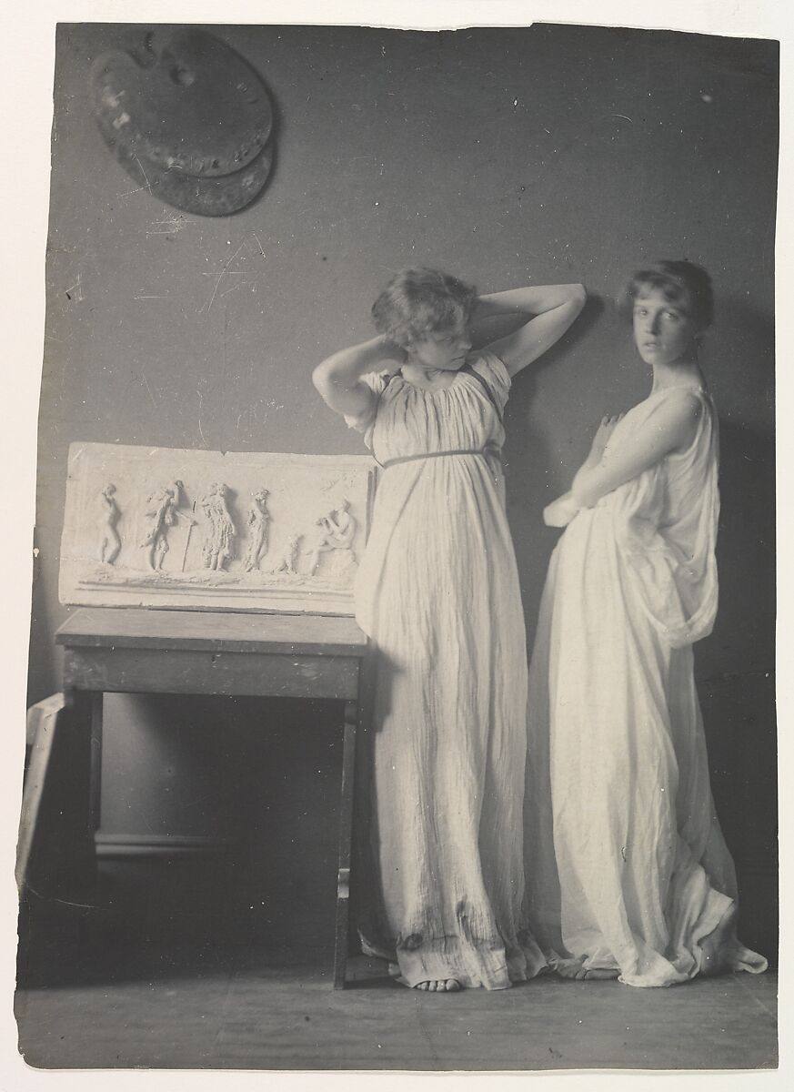 Two young ladies in white Greek Dress, posed side by side, one looking at the camera while the other looks off to the right. 