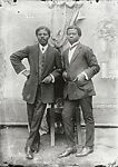 Two dapper young men leaning on a chair